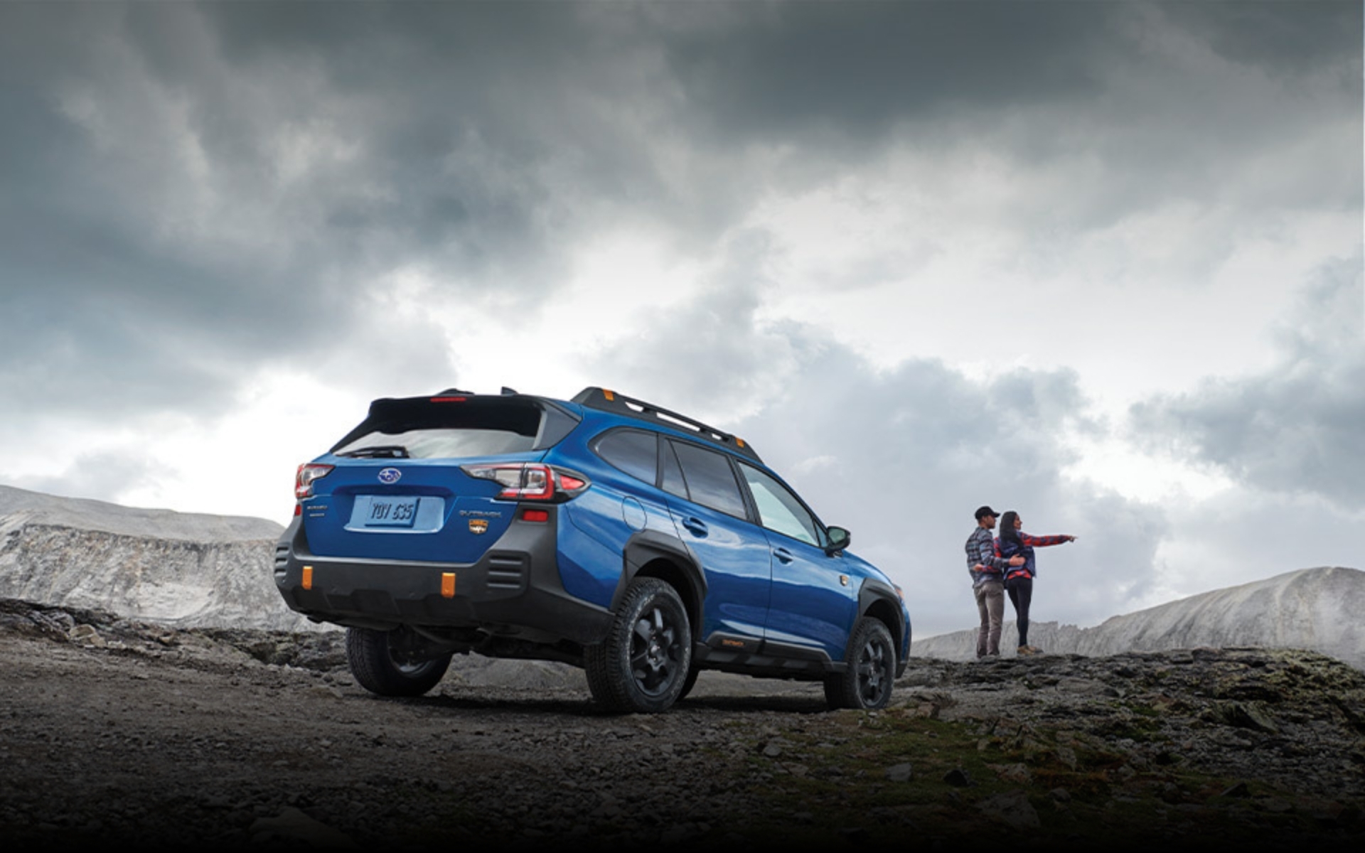 2022 Outback Wilderness in Geyser Blue parked on a rocky ridge behind a couple observing the scenery.